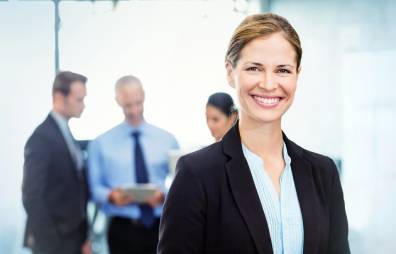 Frau im Business-Look lächelt in die Kamera, dahinter 3 Büro-Mitarbeitende im Gespräch