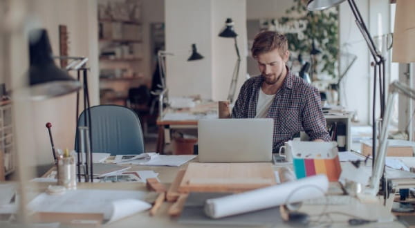 Mann sitzt am vollen Schreibtisch, schaut auf Laptop