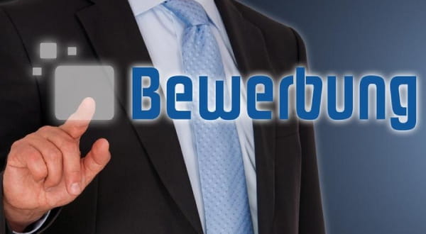 Person im Anzug im Hintergrund, zeigt mit Finger auf die Schrift "BEWERBUNG" im Vordergrund