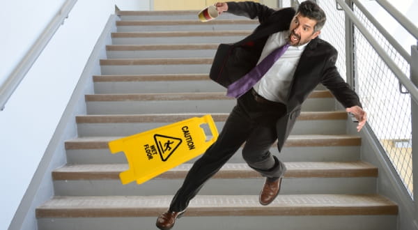 Mann im Anzug auf Treppe am hinfallen, neben einem gelben "Caution - wet floor" Schild.