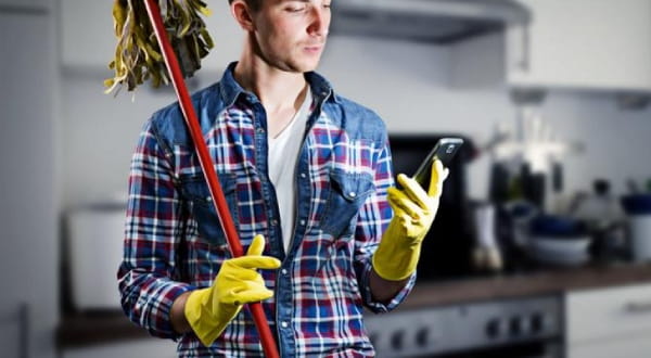 Mann steht mit Putzhandschuhen, hält Wischmopp während er am Handy beschäftigt ist