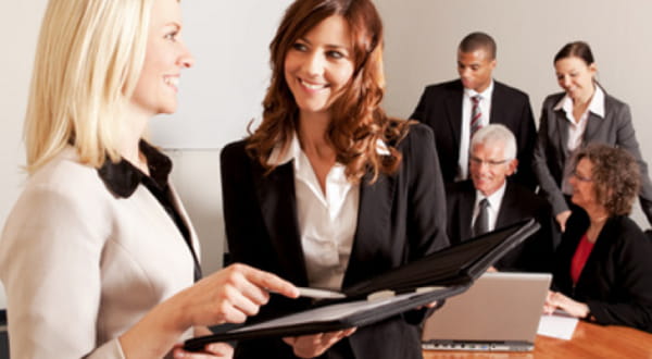 Zwei Frauen in Businesskleidung im Vordergrund lächelnd am Unterhalten, Gruppe von Menschen im Hintergrund