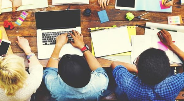 Sicht von oben: Gruppe sitzt an großem Tisch, am Laptop, Block, Tablet...