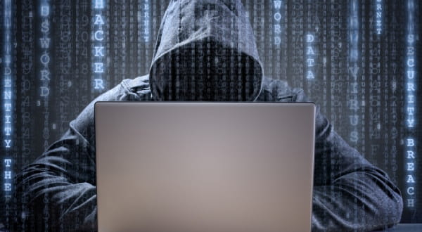 Person mit Kapuze sitzt hinter Laptop, umgeben von codierten Daten
