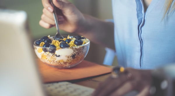 Person sitzt am Tisch, isst mit einer Hand Cornflakes mit Blaubeeren und bedient mit der anderen den laptop