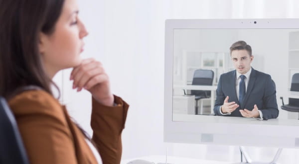 Frau sitzt und schaut auf PC-Display, auf dem ein Mann im Anzug am reden ist