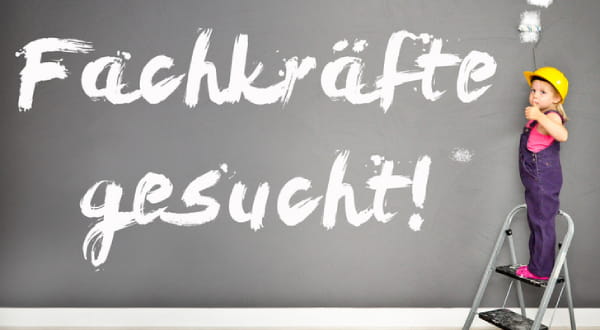 "Fachkraft gesucht!" an Wand gemalt, Kind steht auf Leiter in Malerkleidung und Schutzhelm