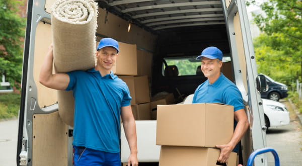 vor Transporter voll mit Kisten stehen zwei Männer, die lächeln in Kamera und tragenTeppich und Kisten 
