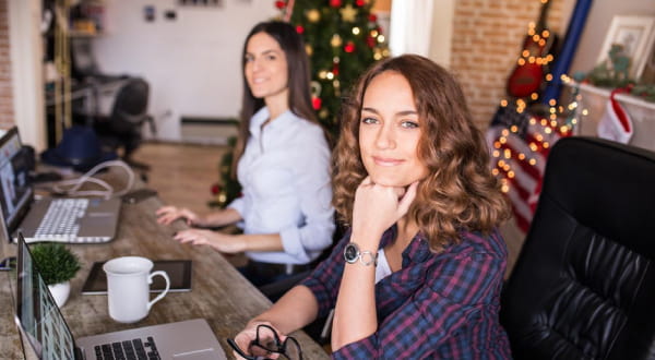 Zwei Frauen sitzen vor Schreibtisch, blicken in die Kamera, Weihnachtsdekoration im Hintergrund